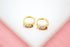 A pair of gold huggie hoop earrings embedded with colorful multi-gemstones rests on a white display surface against a pink background.