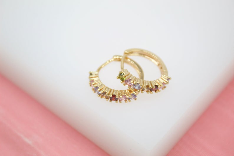 A pair of gold hoop earrings features vibrant multicolored gemstones set along the bands against a soft white and pink background.
