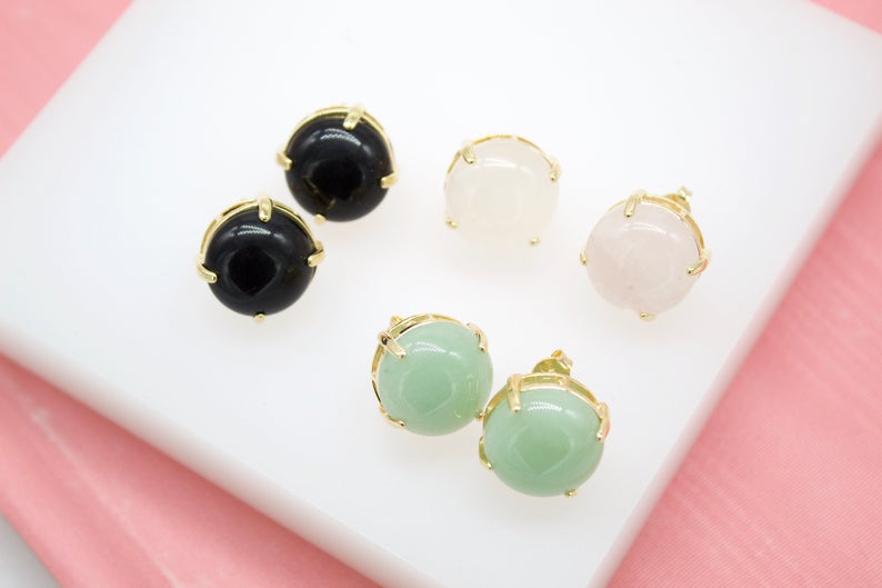 Three pairs of round gemstone stud earrings in black, white, and green with gold prong settings rest on a white display surface.