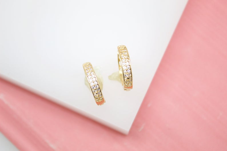 A pair of gold huggie hoop earrings encrusted with sparkling cubic zirconia stones rests elegantly on a white and pink surface.