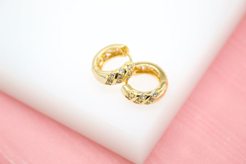 A pair of gold huggie hoop earrings embedded with sparkling cubic zirconia stones rests on a white display block.