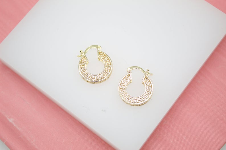 Two gold hoop earrings featuring a Greek key design rest on a white display surface against a pink background.