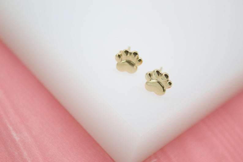 A pair of polished gold paw print stud earrings rests on a white display surface against a pink background.