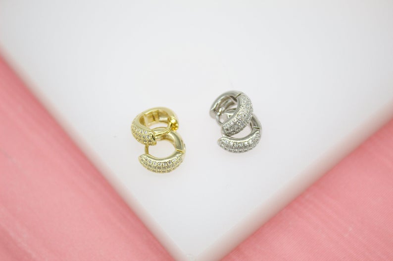 Two pairs of pave-set diamond huggie earrings rest on a white display surface against a pink background, showcasing gold and silver metal finishes.