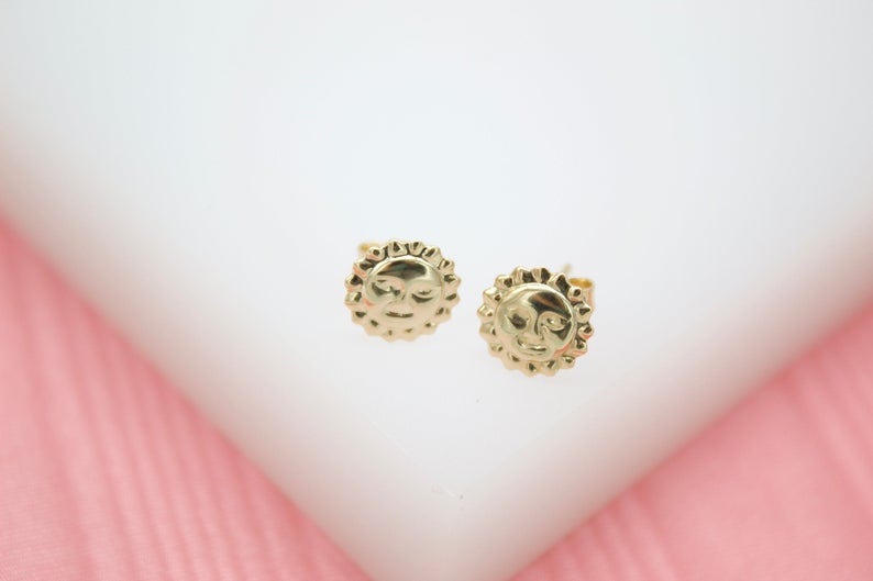 A pair of polished gold sun face stud earrings rests on a white and pink display stand.