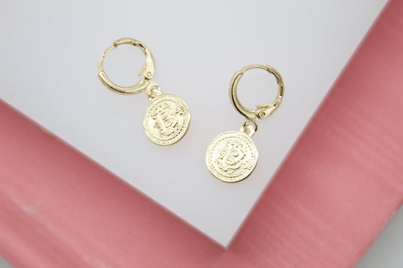 A pair of gold hoop earrings featuring dangling Bitcoin coin charms rests on a white display surface.