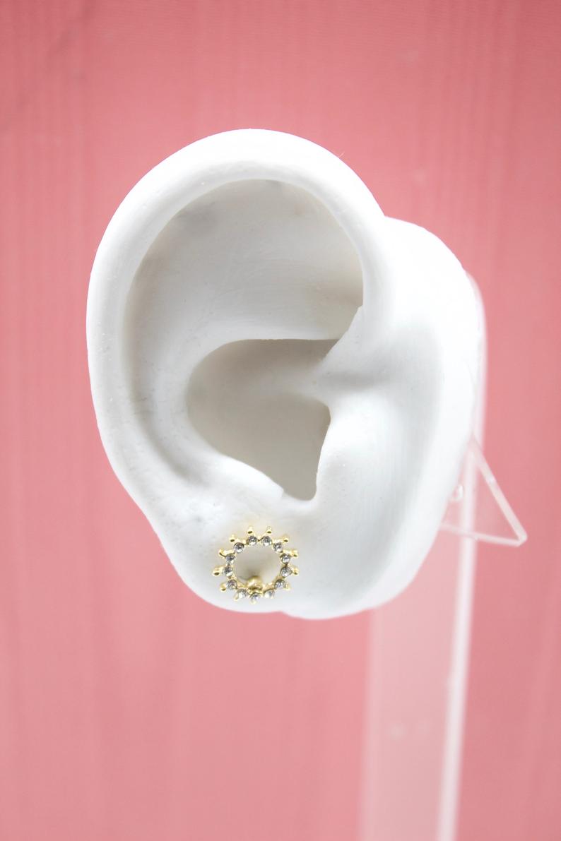 A white mannequin ear displays a sparkling gold sunburst stud earring against a pink background.