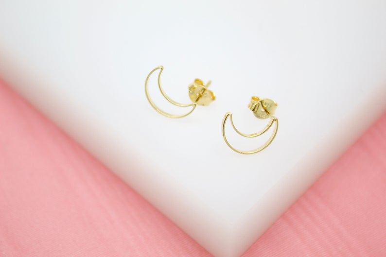 A pair of delicate gold crescent moon stud earrings rests on a white and pink display stand.