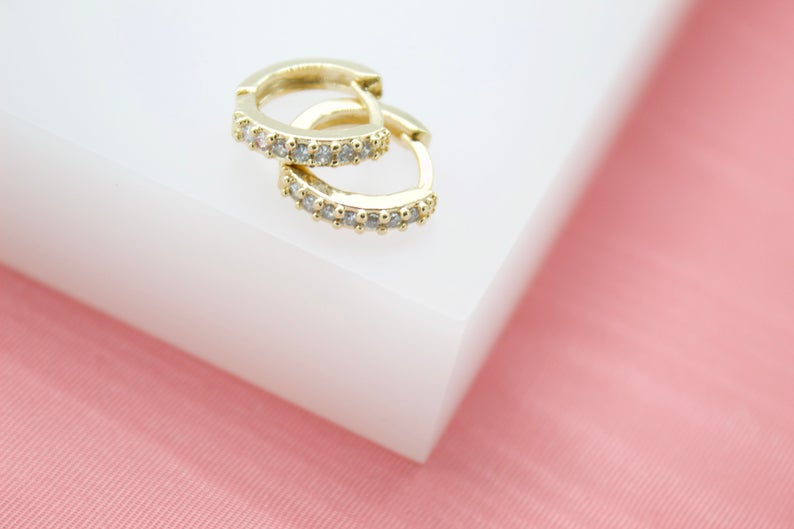 Two delicate gold hoop earrings studded with sparkling cubic zirconia rest on a white display block.