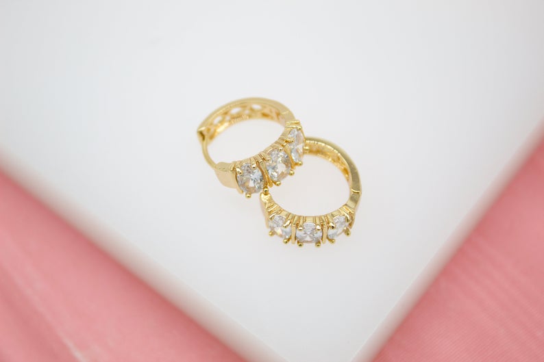 A pair of gold huggie hoop earrings embedded with sparkling cubic zirconia stones rests on a white and pink surface.