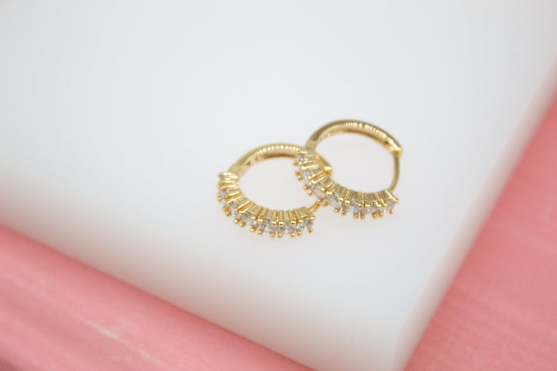 A pair of shimmering gold hoop earrings set with sparkling cubic zirconia stones rests against a soft white and pink background.