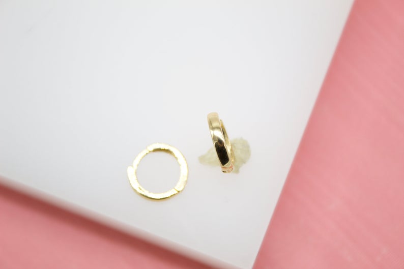A pair of small gold hoop earrings rests on a white display surface against a pink background.