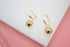 A pair of shiny gold ball leverback earrings rests on a white and pink display.