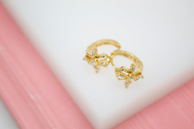 A pair of gold huggie hoop earrings featuring sparkling crystal-emborted bows rest on a white display surface.