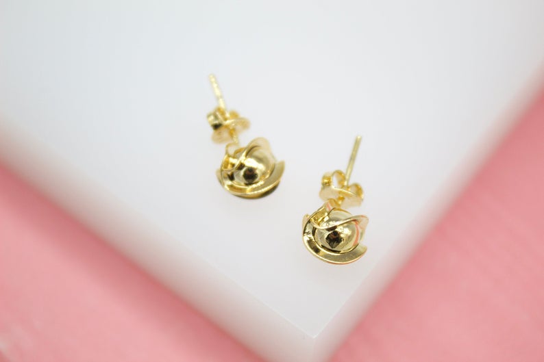 A pair of shiny gold stud earrings rest on a white display surface against a soft pink background.
