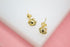 A pair of shiny gold stud earrings rest on a white display surface against a soft pink background.