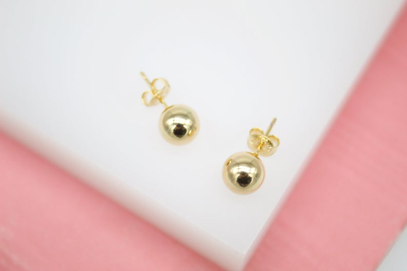 A pair of polished gold ball stud earrings rests against a white and pink background.