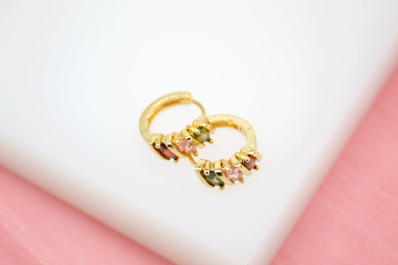 A pair of gold huggie hoop earrings featuring colorful multi-gemstone charms rests on a white and pink surface.