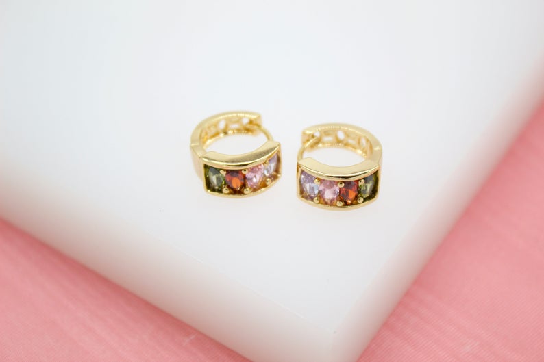 A pair of gold huggie hoop earrings featuring sparkling multicolored gemstones sits on a white surface against a pink background.