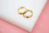 Two gold huggie hoop earrings featuring multicolored gemstones rest on a white surface against a pink background.