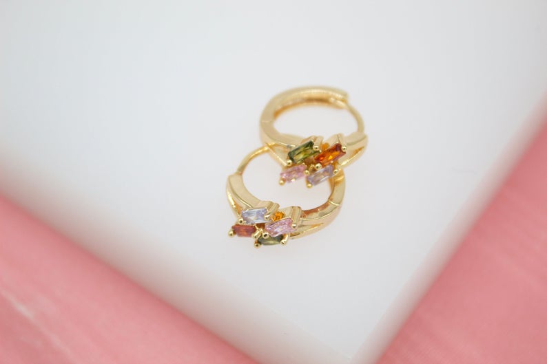 A pair of gold huggie hoop earrings decorated with colorful baguette cubic zirconia stones rests on a white display surface against a soft pink background.