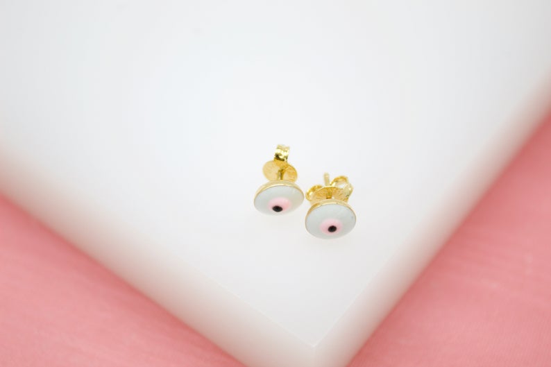 A pair of gold stud earrings featuring a delicate white and pink evil eye design rests on a white display surface.