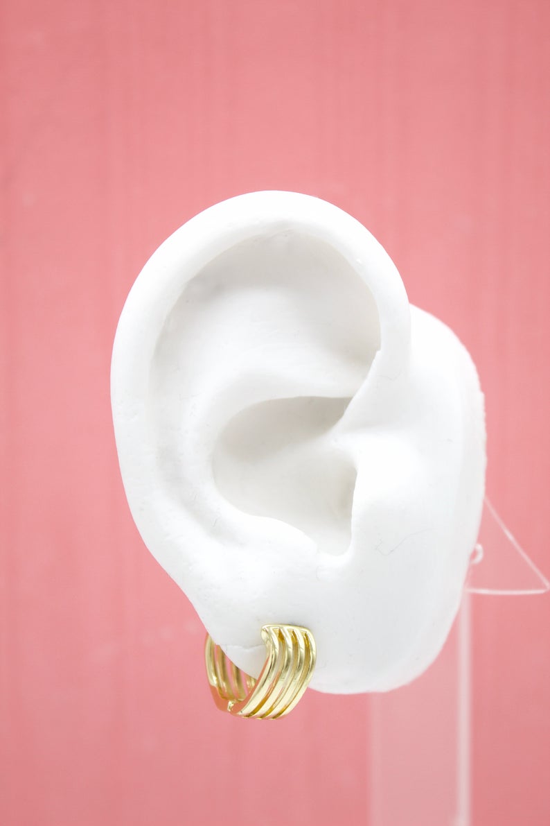 A white mannequin ear displays a stylish multi-band gold hoop earring against a soft pink background.