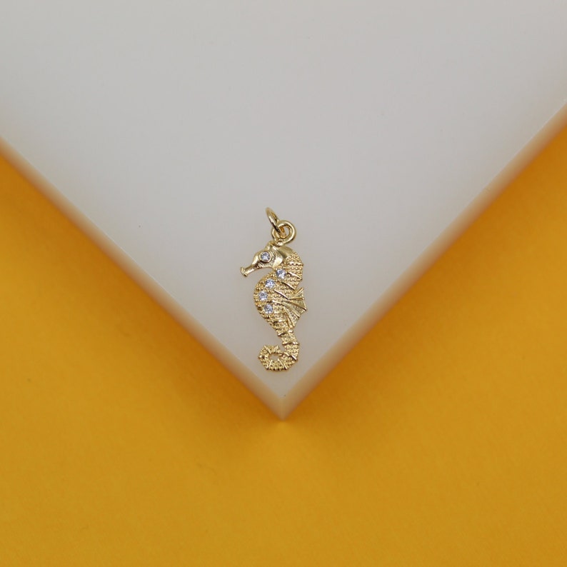 A gold seahorse pendant with sparkling cubic zirconia stones rests on a white display surface against a bright yellow background.