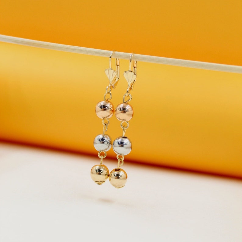 A pair of tri-color gold drop earrings featuring alternating rose, white, and yellow gold beads hangs from a thin wooden stick against a warm yellow and white backdrop.