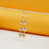 A pair of tri-color gold drop earrings featuring alternating rose, white, and yellow gold beads hangs from a thin wooden stick against a warm yellow and white backdrop.
