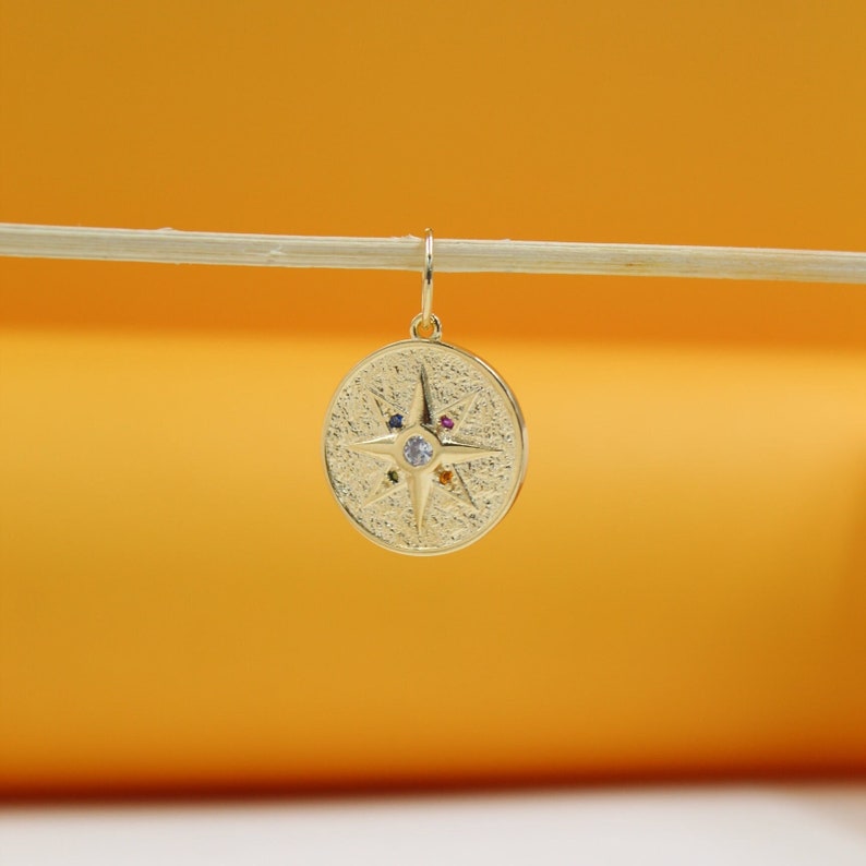 A textured gold compass pendant with colorful gemstone accents hangs from a thin wooden stick against an orange background.