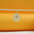 A textured gold compass pendant with colorful gemstone accents hangs from a thin wooden stick against an orange background.
