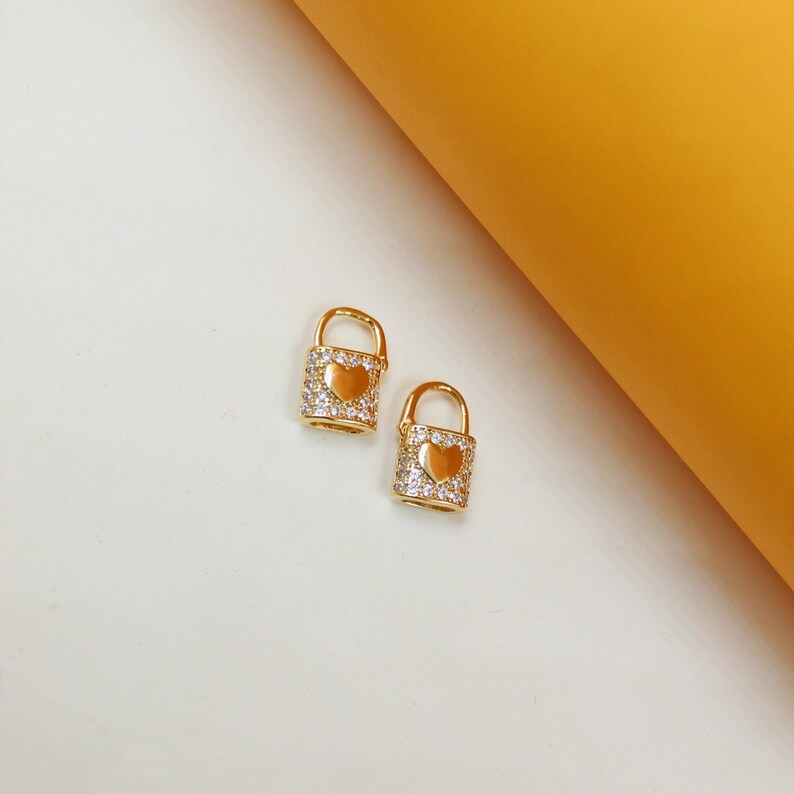 Two gold padlock charms encrusted with cubic zirconia stones and featuring a central heart detail rest on a white surface next to a yellow background.