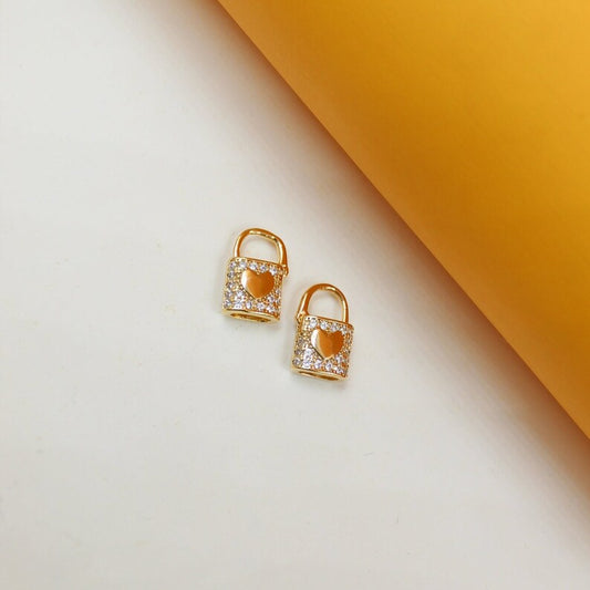 Two gold padlock charms encrusted with cubic zirconia stones and featuring a central heart detail rest on a white surface next to a yellow background.
