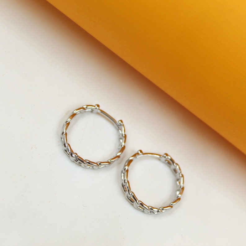 Two polished silver chain-link hoop earrings rest against a clean white and yellow background.