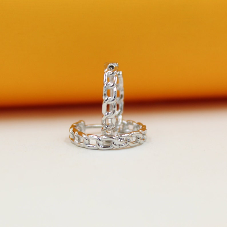 A pair of silver chain link hoop earrings sits upright against a warm orange and white background.