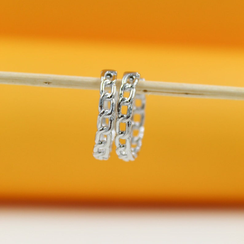 Two silver chain-link hoop earrings hang from a wooden stick against a bright orange background.
