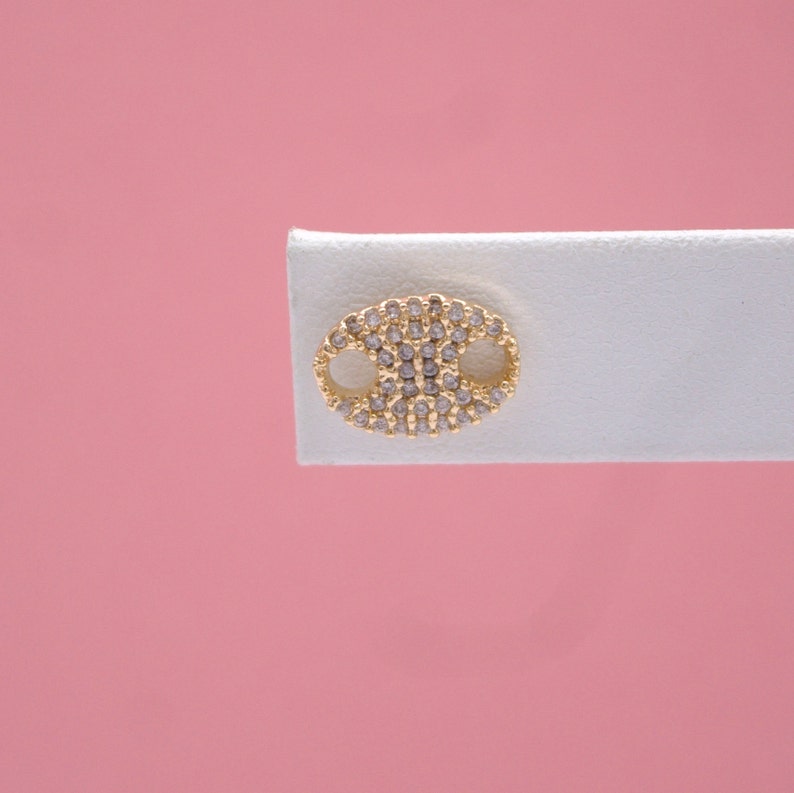 A gold mariner link stud earring encrusted with small clear cubic zirconia stones is displayed on a white jewelry card against a solid pink background.