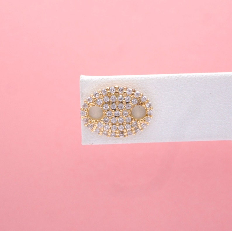 A gold anchor link stud earring encrusted with sparkling cubic zirconia stones is displayed on a white card against a soft pink background.
