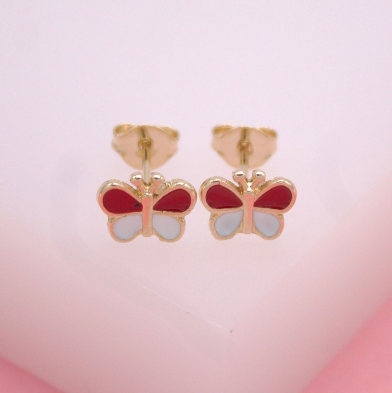 A pair of gold butterfly stud earrings features red and white enamel wings displayed against a soft pink background.