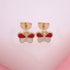 A pair of gold butterfly stud earrings features red and white enamel wings displayed against a soft pink background.