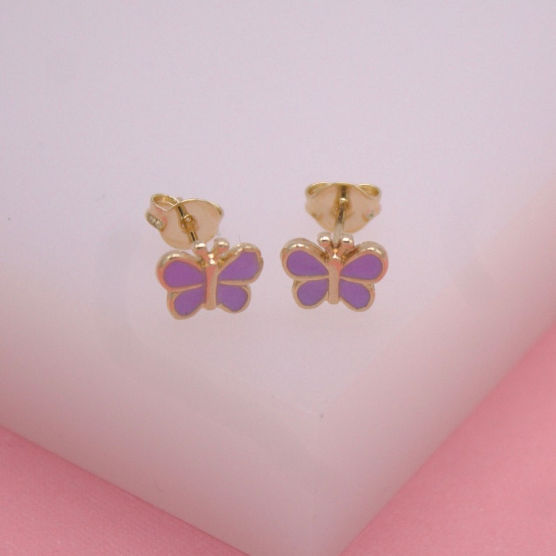 A pair of gold butterfly stud earrings feature vibrant purple enamel wings against a light pink backdrop.