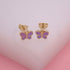 A pair of gold butterfly stud earrings feature vibrant purple enamel wings against a light pink backdrop.