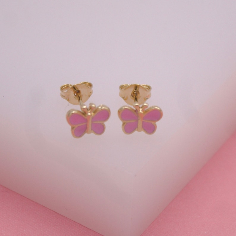 A pair of gold butterfly stud earrings with pink enamel wings rests against a soft pink and white background.