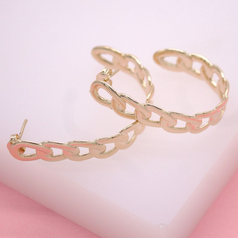 A pair of gold chain link hoop earrings rests on a soft pink surface.