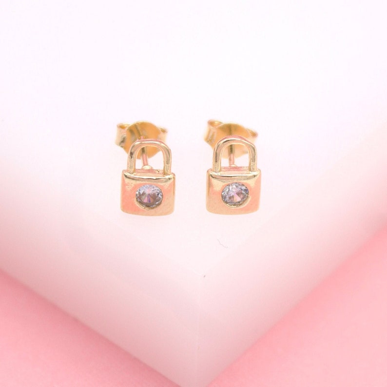A pair of gold padlock stud earrings sparkle with tiny embedded crystals against a soft pink and white background.