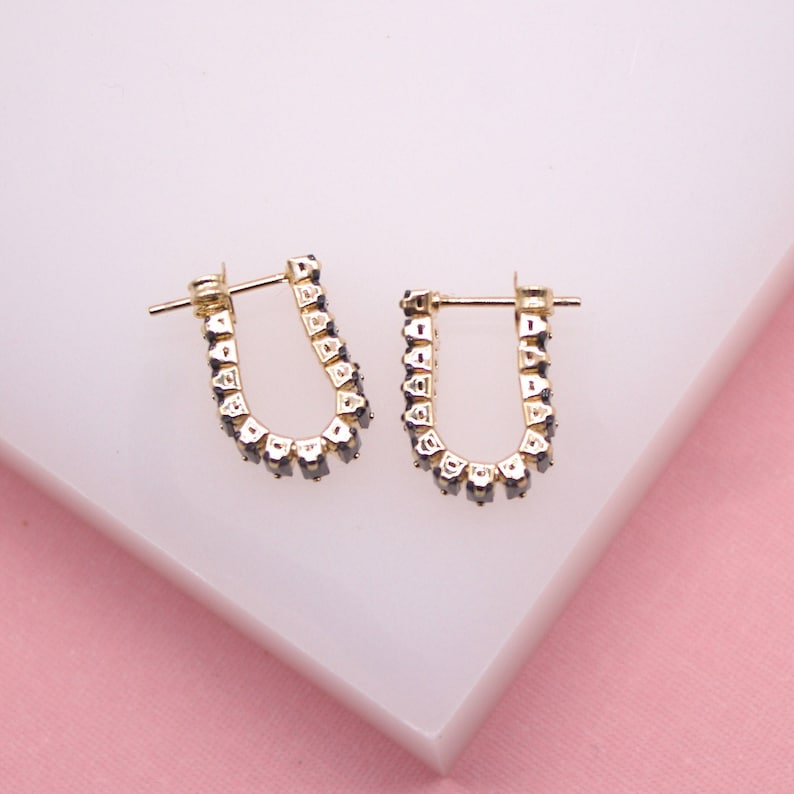 A pair of gold huggie hoop earrings featuring dark rectangular stones rests on a white and pink surface.