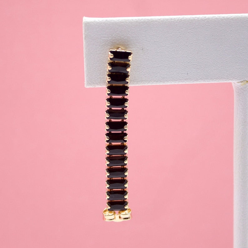 A long gold earring featuring a row of dark red baguette-cut stones hangs against a soft pink background.