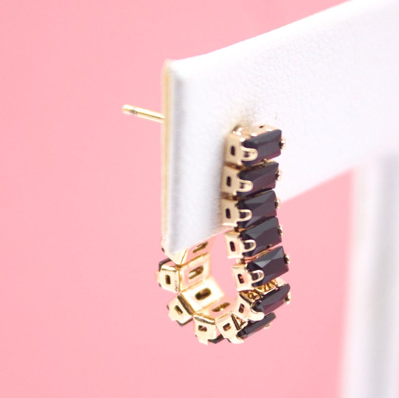 A gold huggie hoop earring featuring a row of dark black baguette-cut stones hangs on a white display card against a soft pink background.