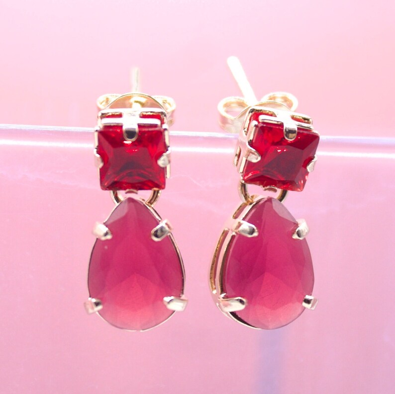 A pair of gold earrings featuring square-cut red stones suspended above larger teardrop-shaped pink gemstones rests against a soft pink backdrop.